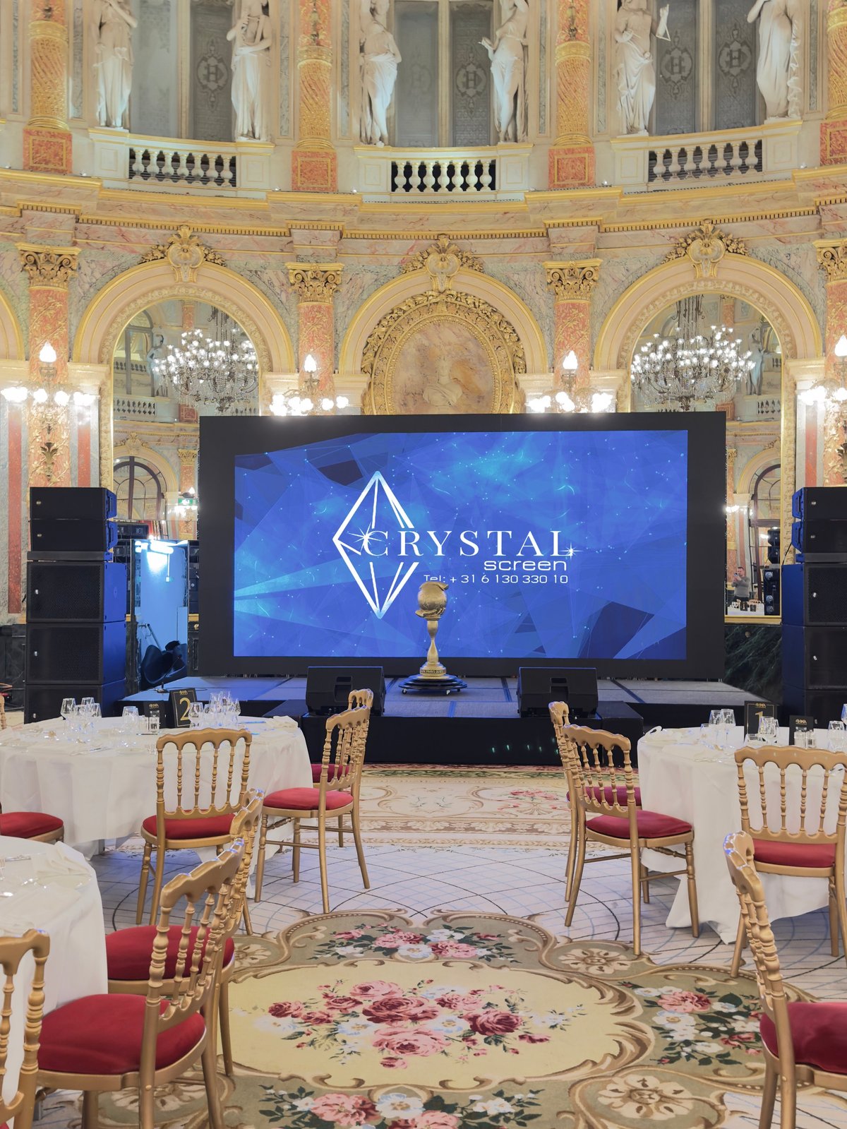 Crystal logo on blue display screen in ornate ballroom with gold chairs, chandeliers, and classical architecture