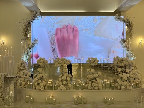 Large-scale window display with white floral arrangements, candles, and a giant pink hand projection on screen