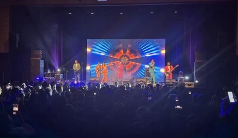 Live concert stage with band performing under colorful blue and red lighting, audience in foreground