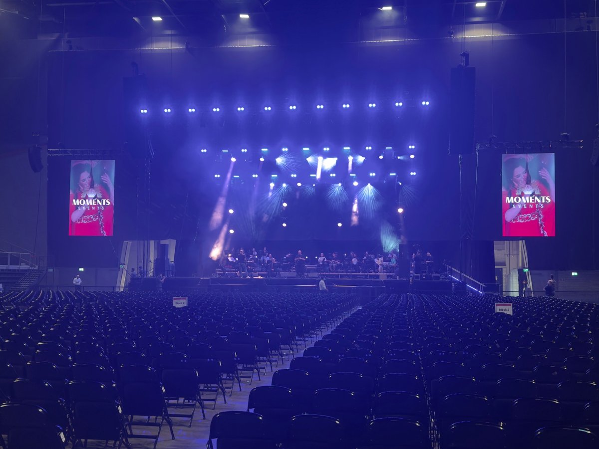 Large concert venue with blue stage lighting, hanging lights, and pink promotional screens displaying a performer on stage