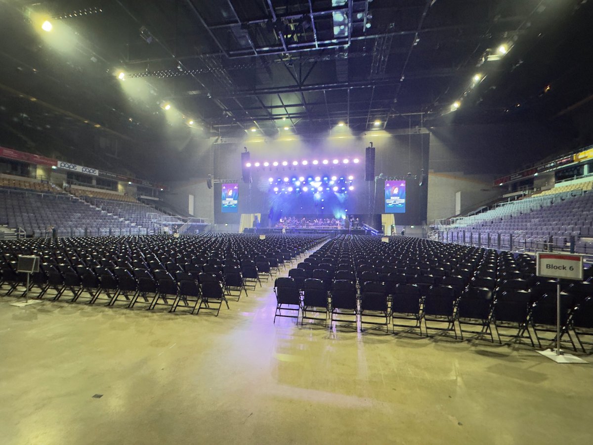 Large indoor arena with empty seating arranged for an event, illuminated stage with blue lighting and spotlights overhead
