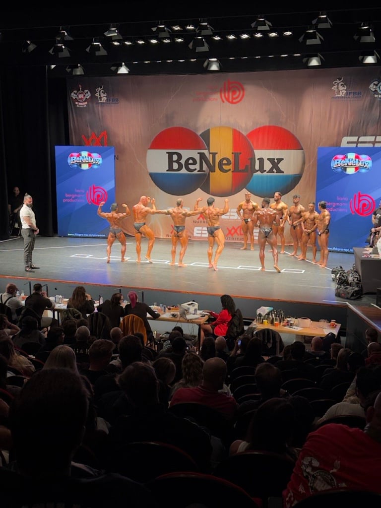 Bodybuilding competition on stage with muscular athletes posing under BeNeLux banner and stage lighting with audience in foreground